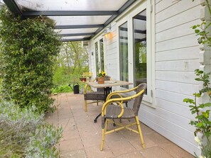 A covered patio area with a table and chairs, surrounded by greenery and a white building. - Holiday Home With 1 Bedroom (Grijpskerke)