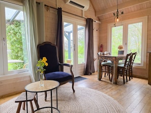 A room with a wooden ceiling, a dining table with chairs, a large window with curtains, and a small round table with a vase of flowers. - Holiday Home With 1 Bedroom (Grijpskerke)