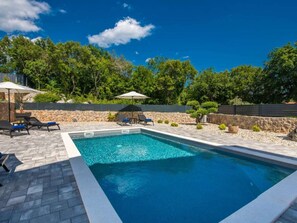 Ein rechteckiger Swimmingpool mit Steinrand, umgeben von Liegestühlen und Sonnenschirmen, vor einer Kulisse aus Bäumen und einer Steinmauer.