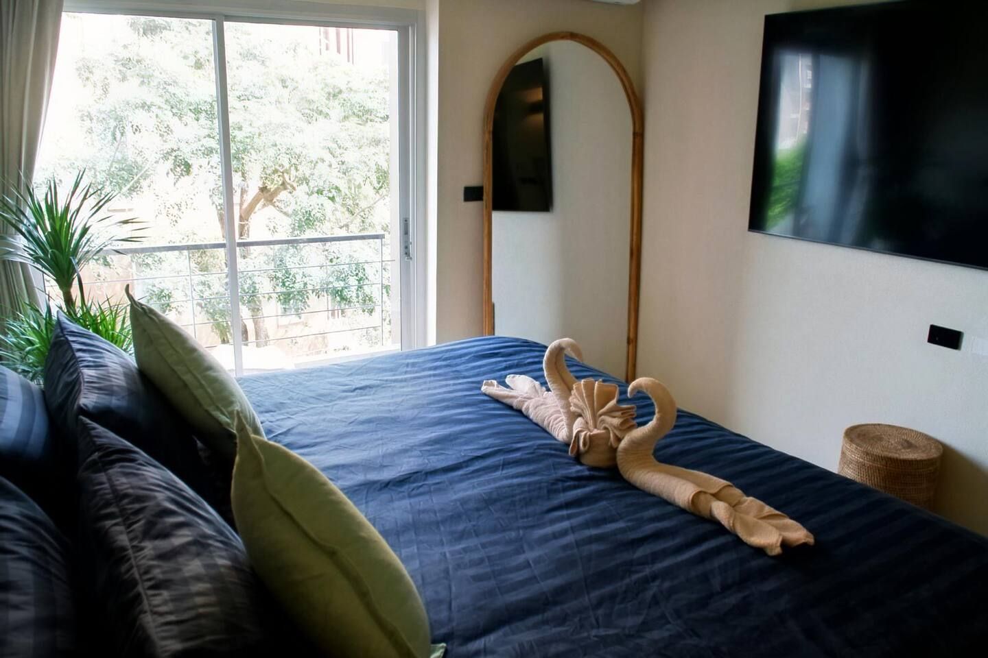 Un lit recouvert d’une couvre-lit bleue, une grande plante en pot, un téléviseur fixé au mur et une fenêtre donnant sur des arbres.