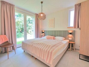 A bedroom with a bed, two bedside tables, a chair, a hanging light fixture, and a window with curtains. - Holiday Home Zone (Callantsoog)