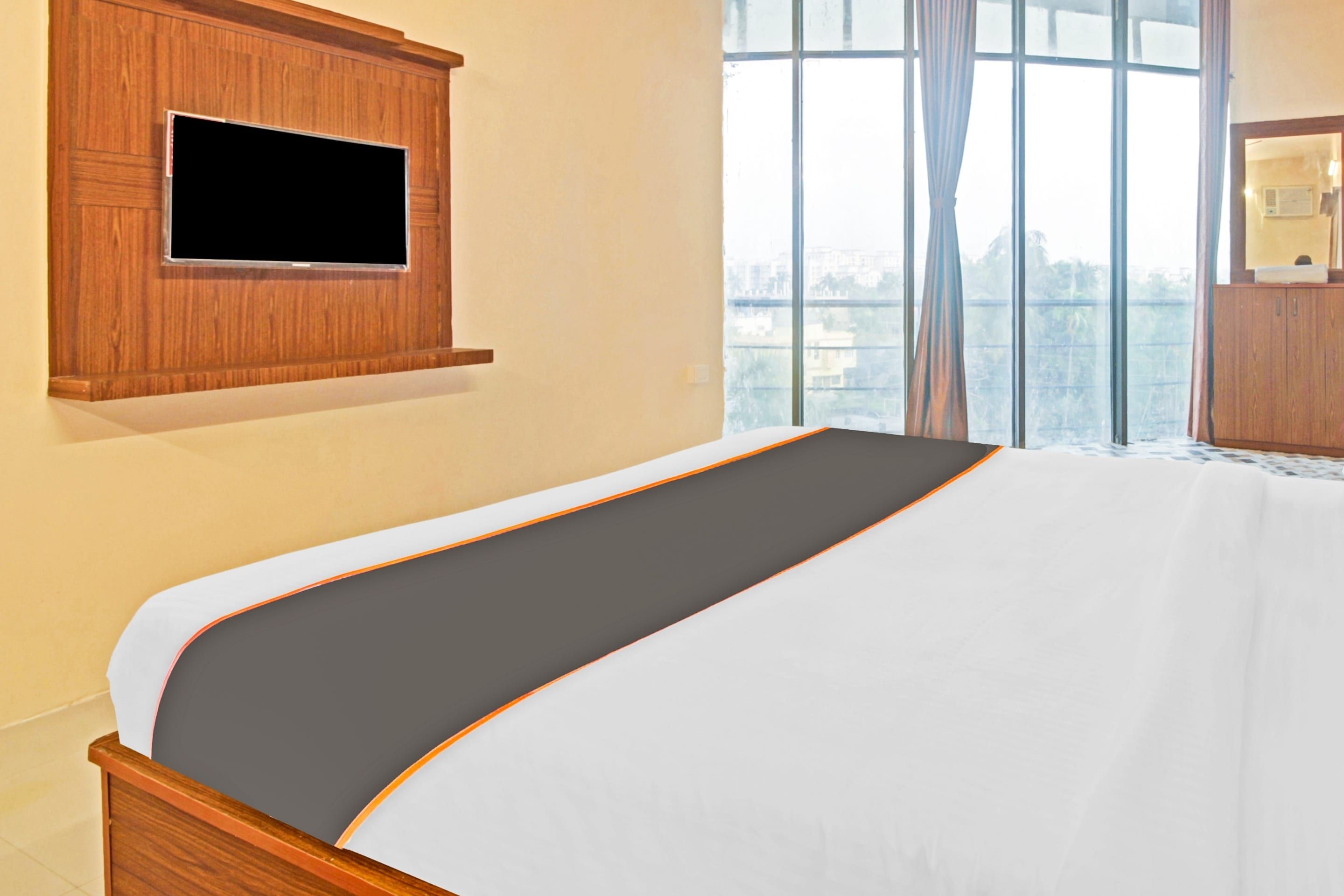 A hotel room with a bed, a television mounted on a wooden panel, and a window with a view of buildings.
