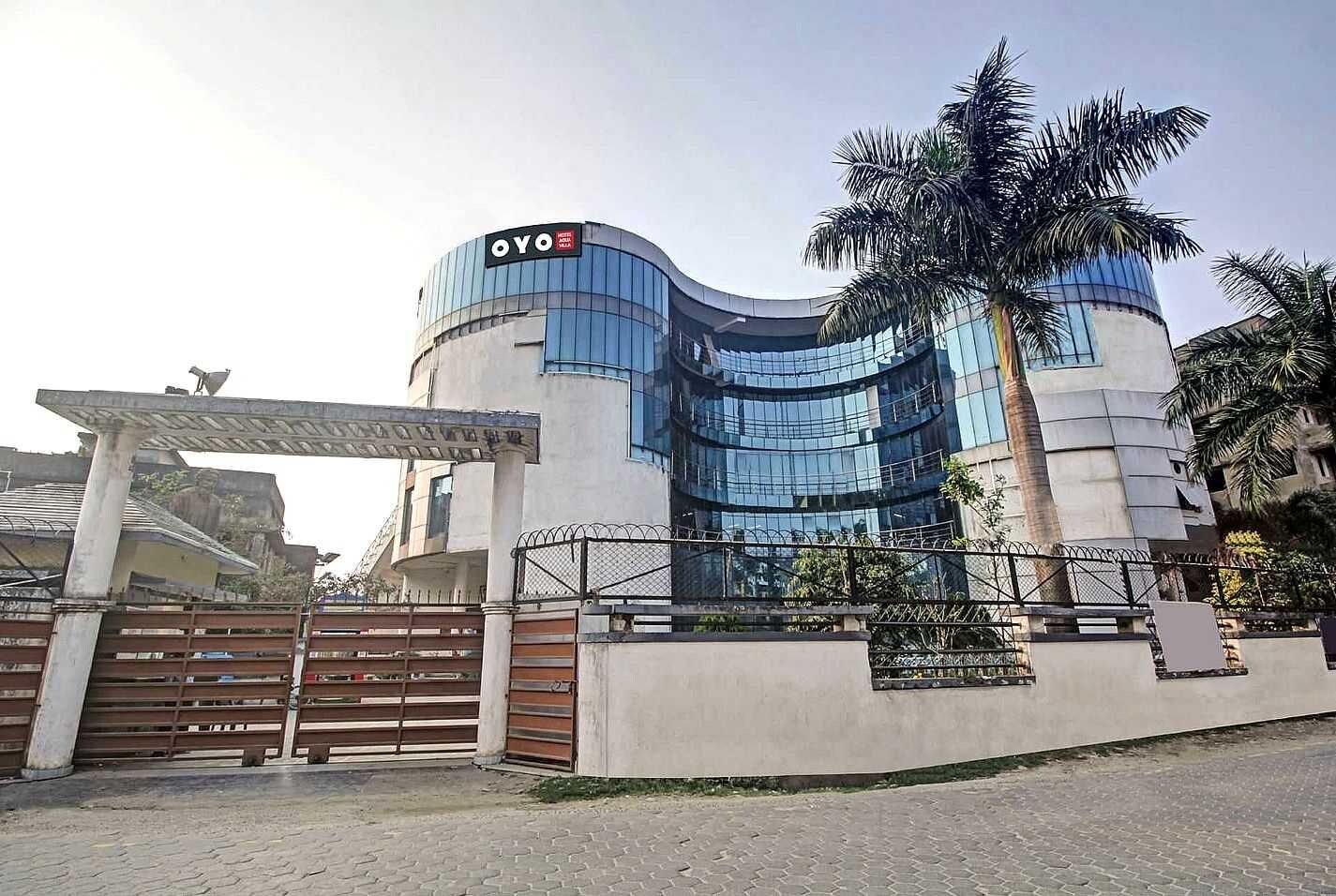 A modern hotel with a curved glass facade, a gated entrance, and a palm tree in the foreground.