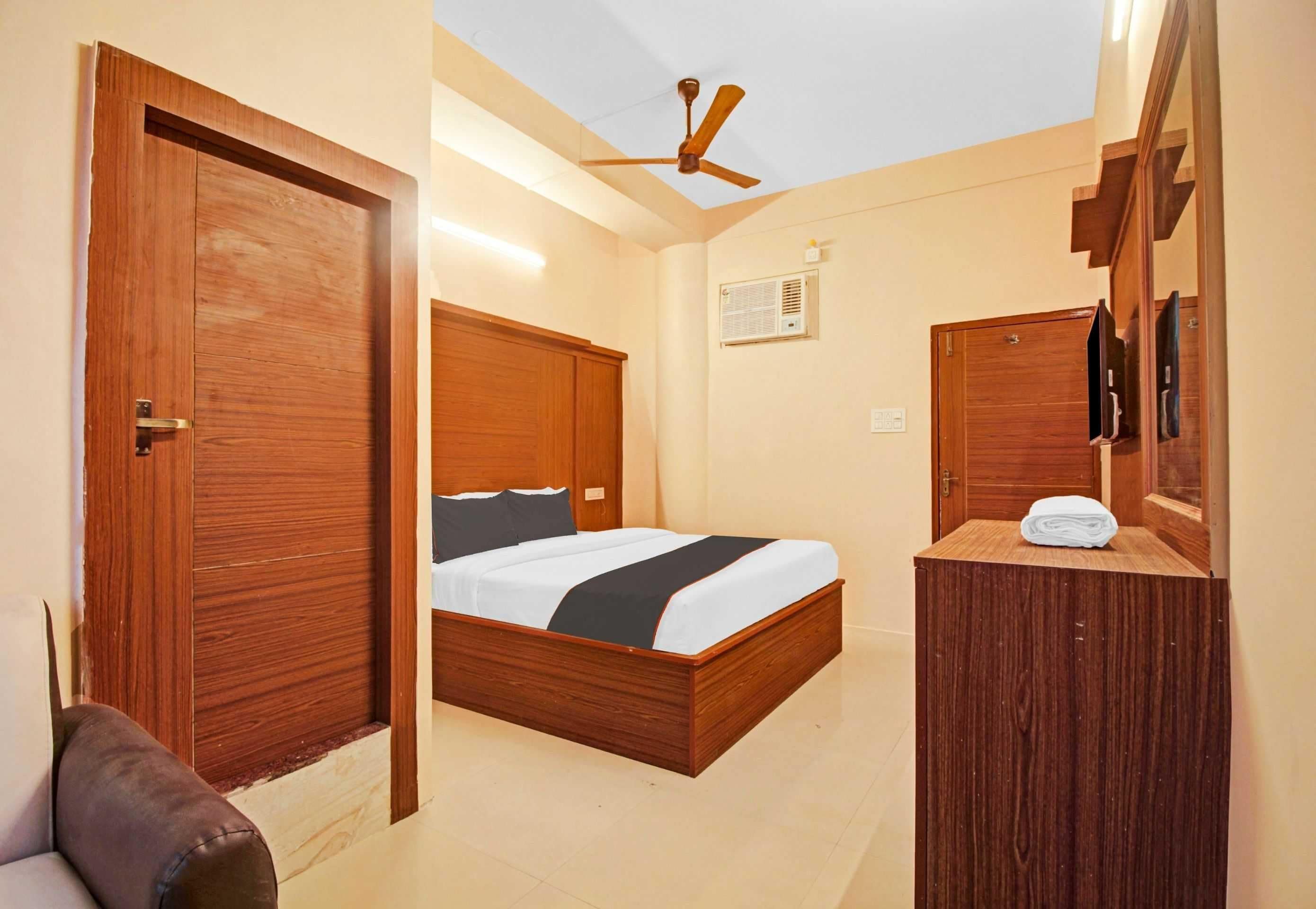 Habitación de hotel con cama y armario de madera, ventilador de techo y ventana pequeña con unidad de aire acondicionado.