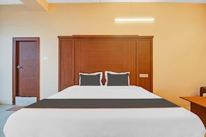 Una habitación de hotel con cabecera de madera, cama con ropa blanca y dos almohadas, mesita de noche y una puerta.