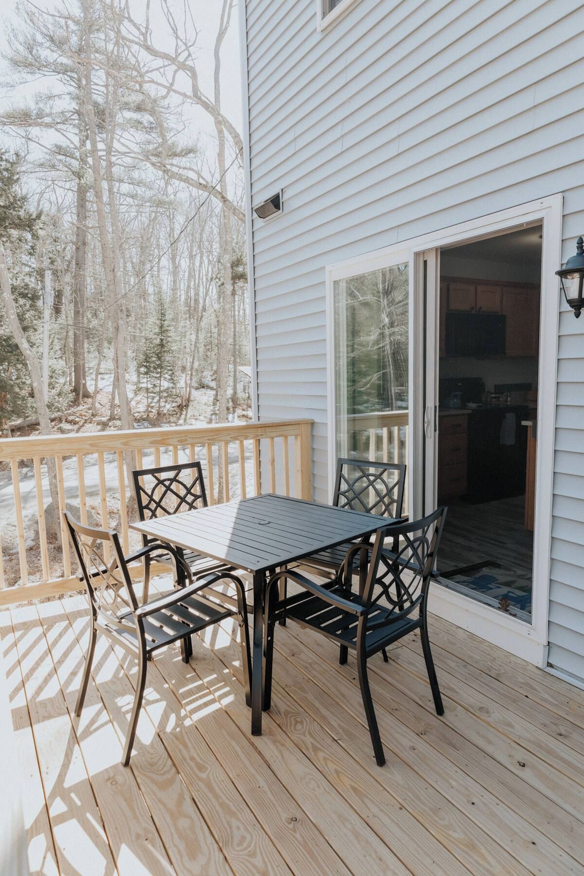 A deck with a table and chairs, a sliding glass door leading to a kitchen, and a view of trees in the background.