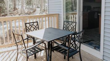 A deck with a table and chairs, a sliding glass door leading to a kitchen, and a view of trees in the background.