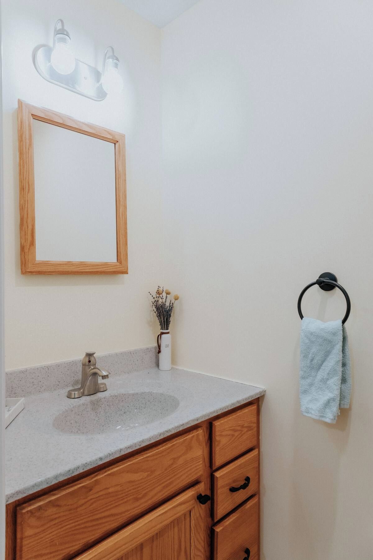 Un baño con tocador de madera, un espejo, aro para toallas y un jarrón con flores secas.