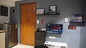 Une pièce avec une porte en bois, un téléviseur à écran plat fixé au mur, une petite étagère avec des photos et un drapeau.