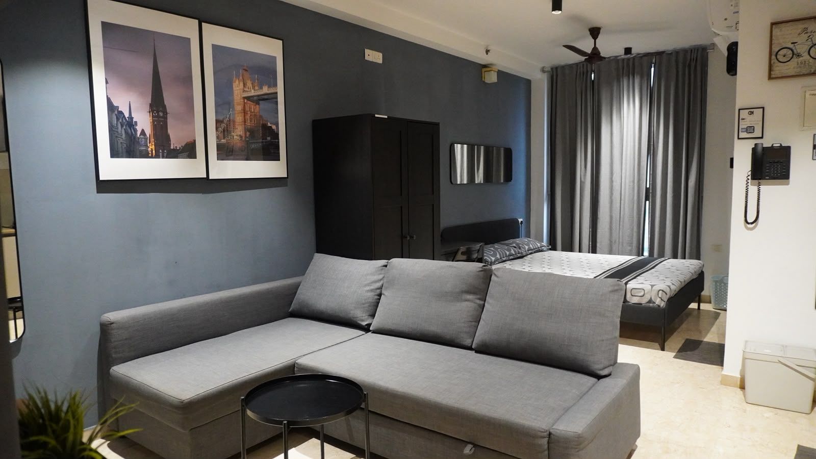 A modern living room with a grey sofa, a small round table, a bed with a patterned bedspread, and a black wardrobe.