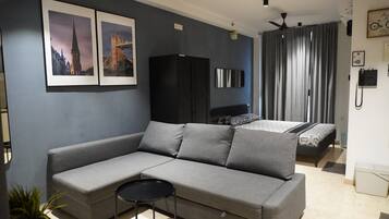 A modern living room with a grey sofa, a small round table, a bed with a patterned bedspread, and a black wardrobe.