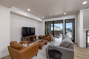 A modern living room with a flat-screen TV, leather sofas, a coffee table, and a large sliding glass door leading to a balcony with a view of buildings and a parking area. - New! Monthly Midtown Bozeman Rental + Dogs Welcome (Bozeman)