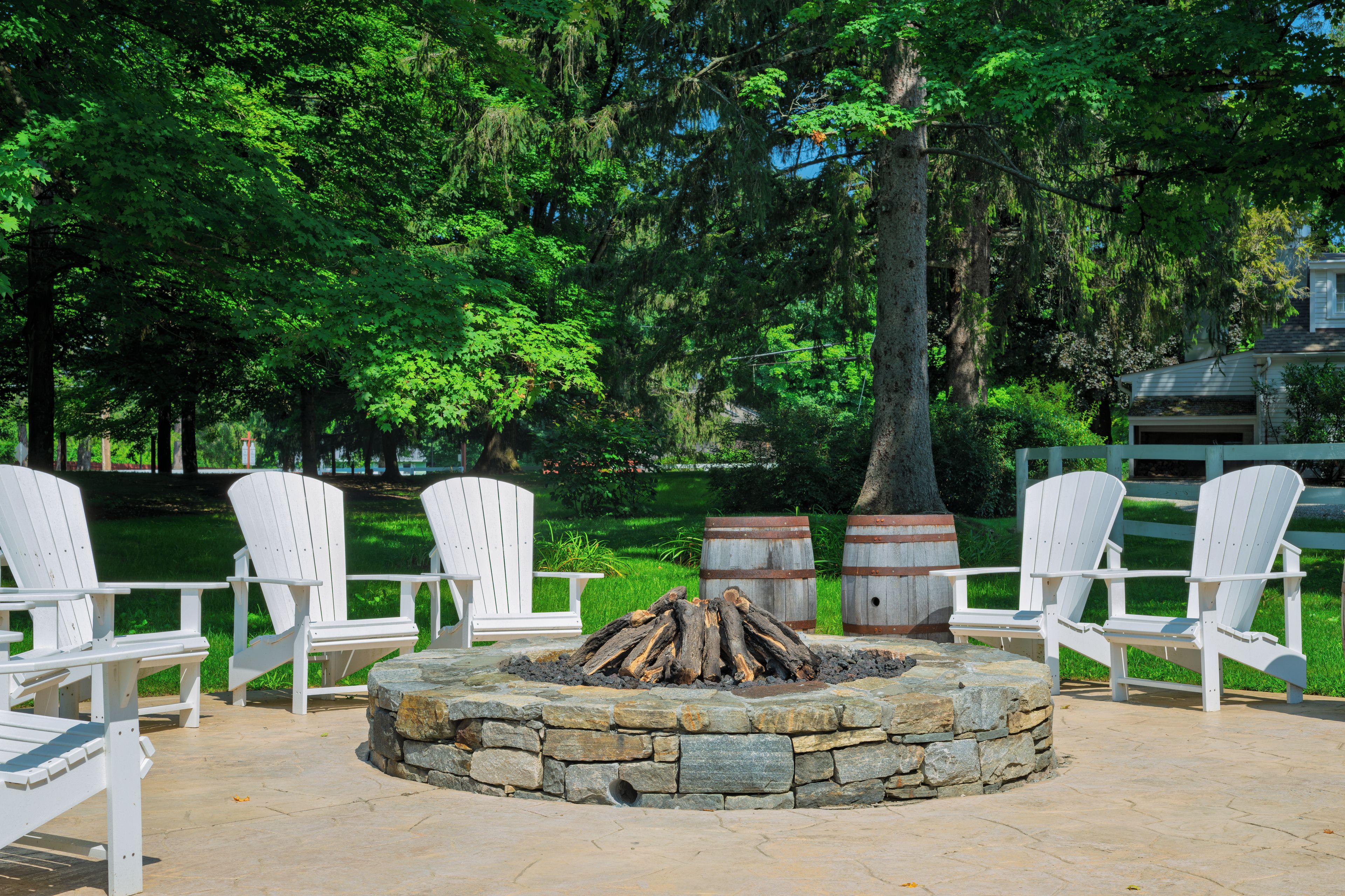 A stone fire pit surrounded by white Adirondack chairs in a park setting.
