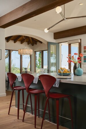 A modern kitchen with a bar area, red bar stools, a hanging light fixture, and a view of the outdoors. - Luxury Estate with Infinity Pool & Panoramic Views of Santa Barbara's Coast & Mountains (Santa Barbara)