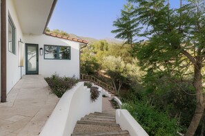 A white building with a tiled entrance, a wooden staircase leading to a door, and a garden with various plants and trees. - Luxury Estate with Infinity Pool & Panoramic Views of Santa Barbara's Coast & Mountains (Santa Barbara)