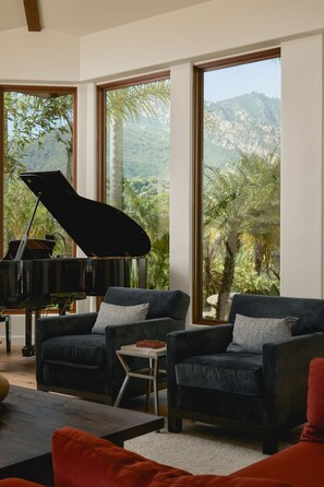 A living room with a grand piano, two dark sofas, and a coffee table. Large windows offer a view of a mountainous landscape. - Luxury Estate with Infinity Pool & Panoramic Views of Santa Barbara's Coast & Mountains (Santa Barbara)