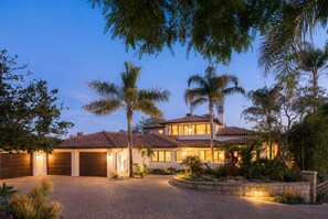 A two-story house with a brick driveway, palm trees, and a well-lit exterior. - Luxury Estate with Infinity Pool & Panoramic Views of Santa Barbara's Coast & Mountains (Santa Barbara)