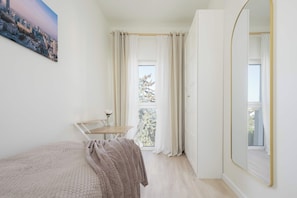 A bedroom with a bed, a small table with a vase, a window with curtains, and a view of trees. - Warsaw | Zbąszyńska 4 | Spacious Apartment | Balcony (Warszawa)