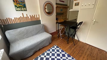 A compact living space with a sofa, a small dining table, a foldable chair, a checkered rug, a mirror, and a kitchen area.
