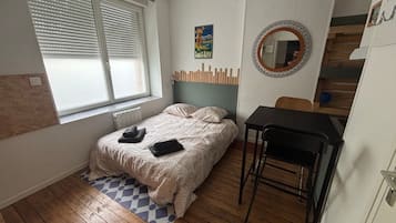 A bedroom with a bed, a small table, a chair, a window with blinds, a radiator, and a poster on the wall.