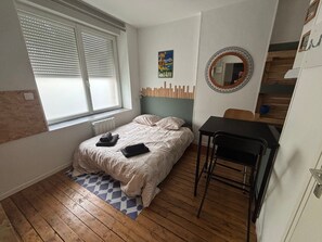 A bedroom with a bed, a small table, a chair, a window with blinds, a radiator, and a poster on the wall. - The Little Golfer - 17m² studio Arras city center (Arras)
