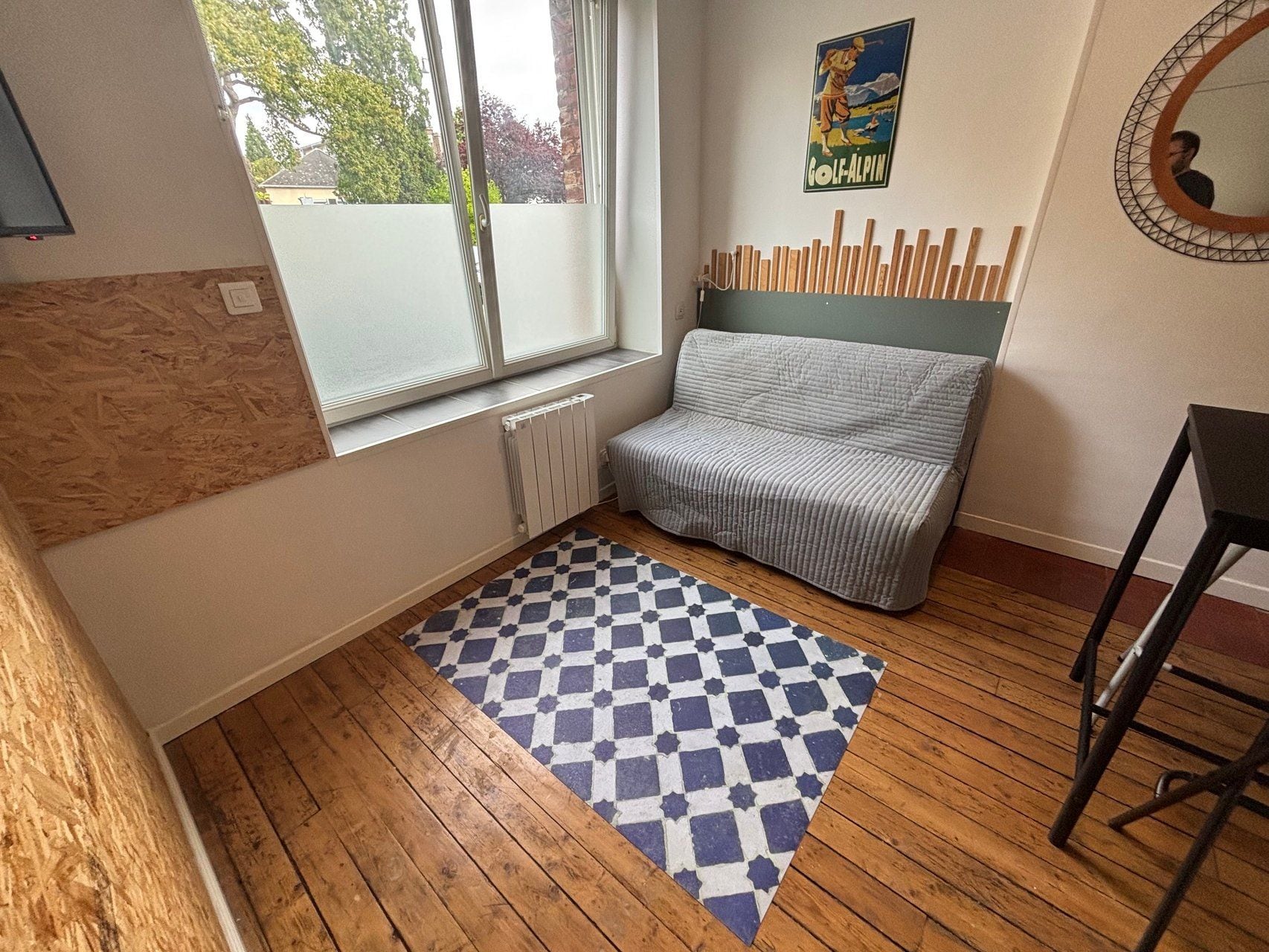 A room with a window, a radiator, a patterned rug, a chair, and a wall-mounted mirror.