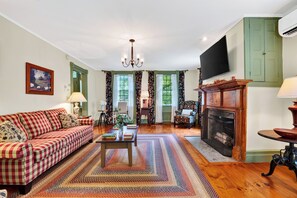 A living room with a checkered sofa, a fireplace, and a television mounted on the wall. - Library + Sunroom + Dining for 8 | Ensuite Baths + Gardens | Preston House (74) (Dover Plains)