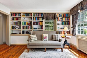 A cozy reading nook with a sofa, a small table, and a bookshelf filled with books. - Library + Sunroom + Dining for 8 | Ensuite Baths + Gardens | Preston House (74) (Dover Plains)