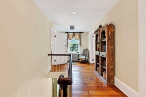 A hallway with a wooden floor, a chair, a bookshelf, and a window with curtains. - Library + Sunroom + Dining for 8 | Ensuite Baths + Gardens | Preston House (74) (Dover Plains)