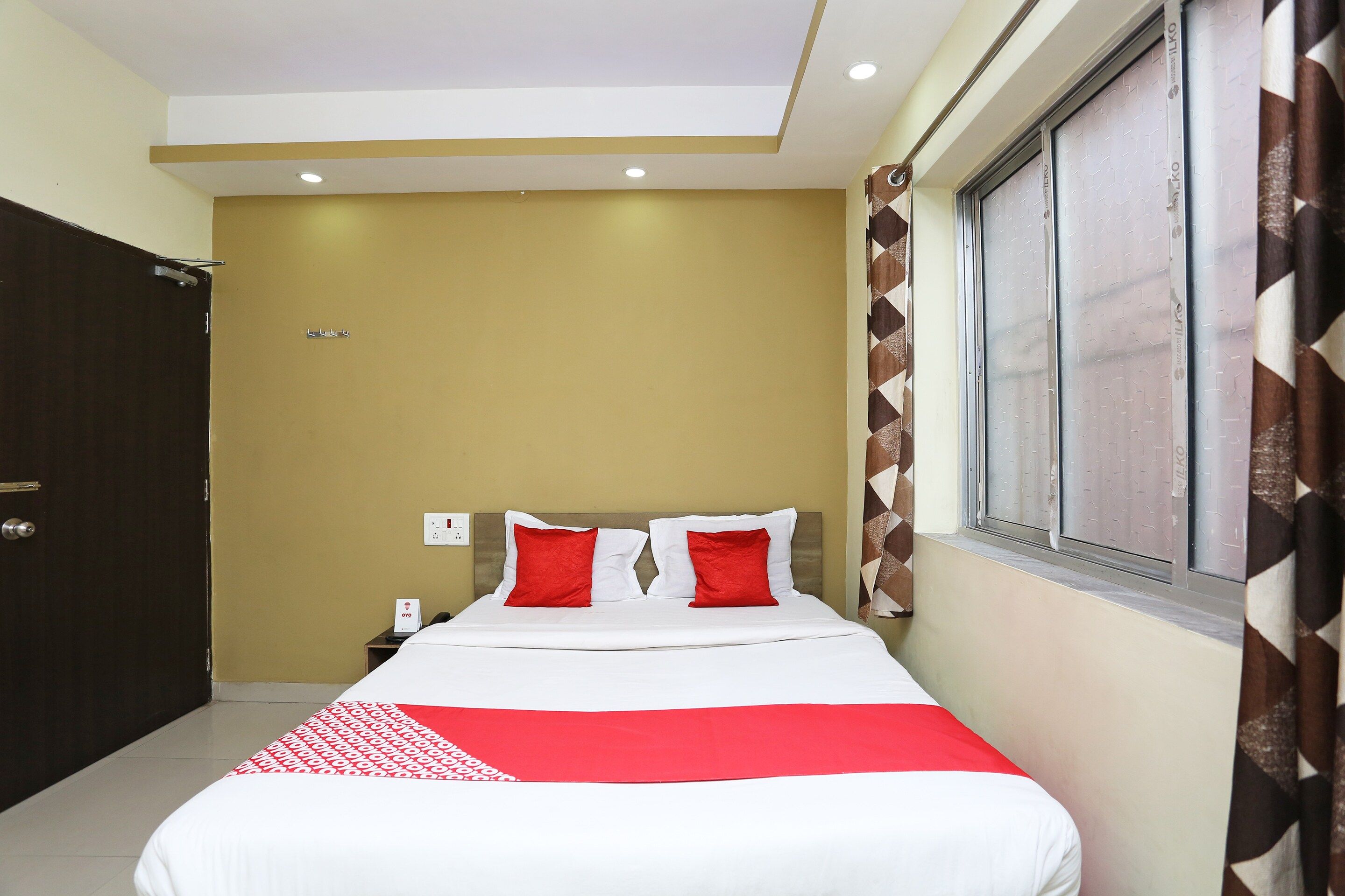 Una habitación de hotel con cama, mesita de noche, ventana con cortinas estampadas y una puerta.