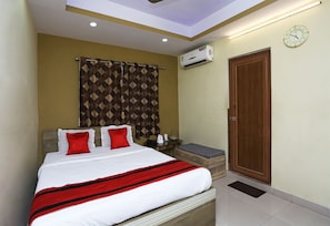 A hotel room with a bed, a bedside table, an air conditioning unit, a wooden door, and a clock on the wall.