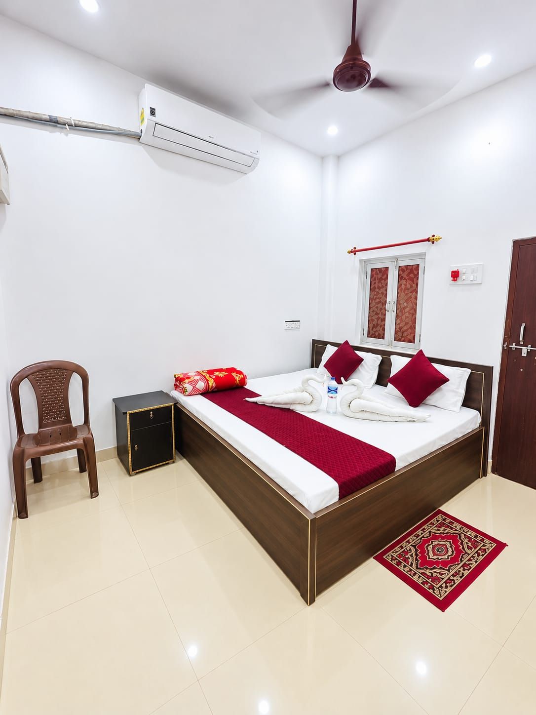 Quarto de hotel com cama, uma cadeira, uma mesinha, aparelho de ar condicionado, ventoinha no teto e tapete vermelho estampado.