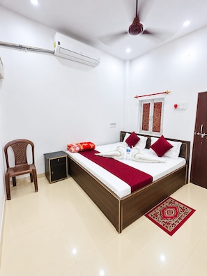 Habitación de hotel con cama, una silla, una mesita, unidad de aire acondicionado, ventilador de techo y alfombra roja estampada.