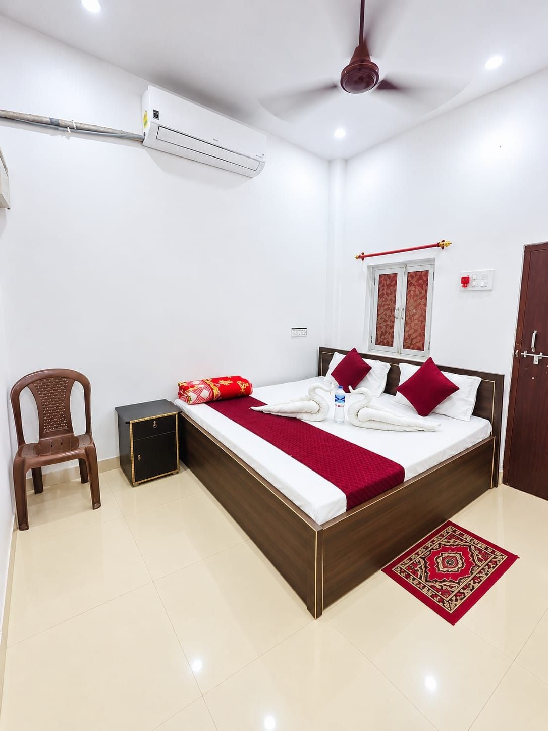 Une chambre d’hôtel avec un lit, une chaise, une petite table, un appareil de climatisation, un ventilateur de plafond et un tapis rouge à motifs.