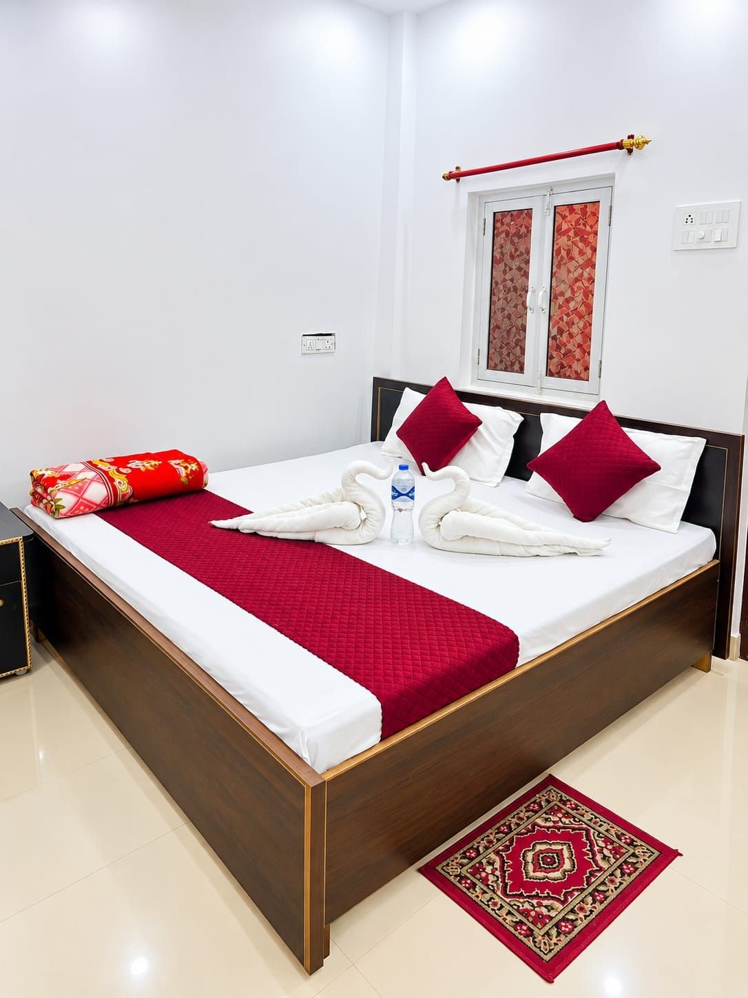 Une chambre d’hôtel avec un lit, une couverture rouge, une bouteille d’eau et un oreiller rouge.