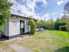A white brick house with a black door, number 413, a small porch, and a well-maintained lawn with outdoor furniture. - Boshuisje Juniper (De Koog)