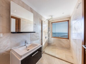 Ein modernes Badezimmer mit Marmorboden, einem weißen Waschbecken, einem Spiegel, einer Duschkabine und einem Fenster mit Blick.