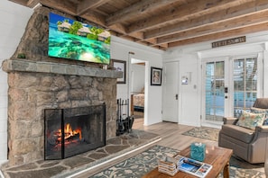 A stone fireplace with a TV mounted above it, a cozy seating area with a leather sofa, and a view of the outdoors through a glass door. - Classic Mountain Cottage | Near Lake & SkyPark + Trails | Green Gables Cottage (Lake Arrowhead)