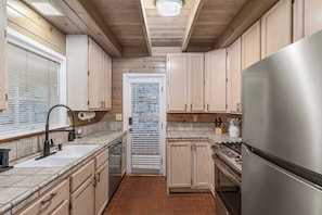 A kitchen with wooden cabinets, a stainless steel refrigerator, a tiled backsplash, and a window with blinds. - Classic Mountain Cottage | Near Lake & SkyPark + Trails | Green Gables Cottage (Lake Arrowhead)