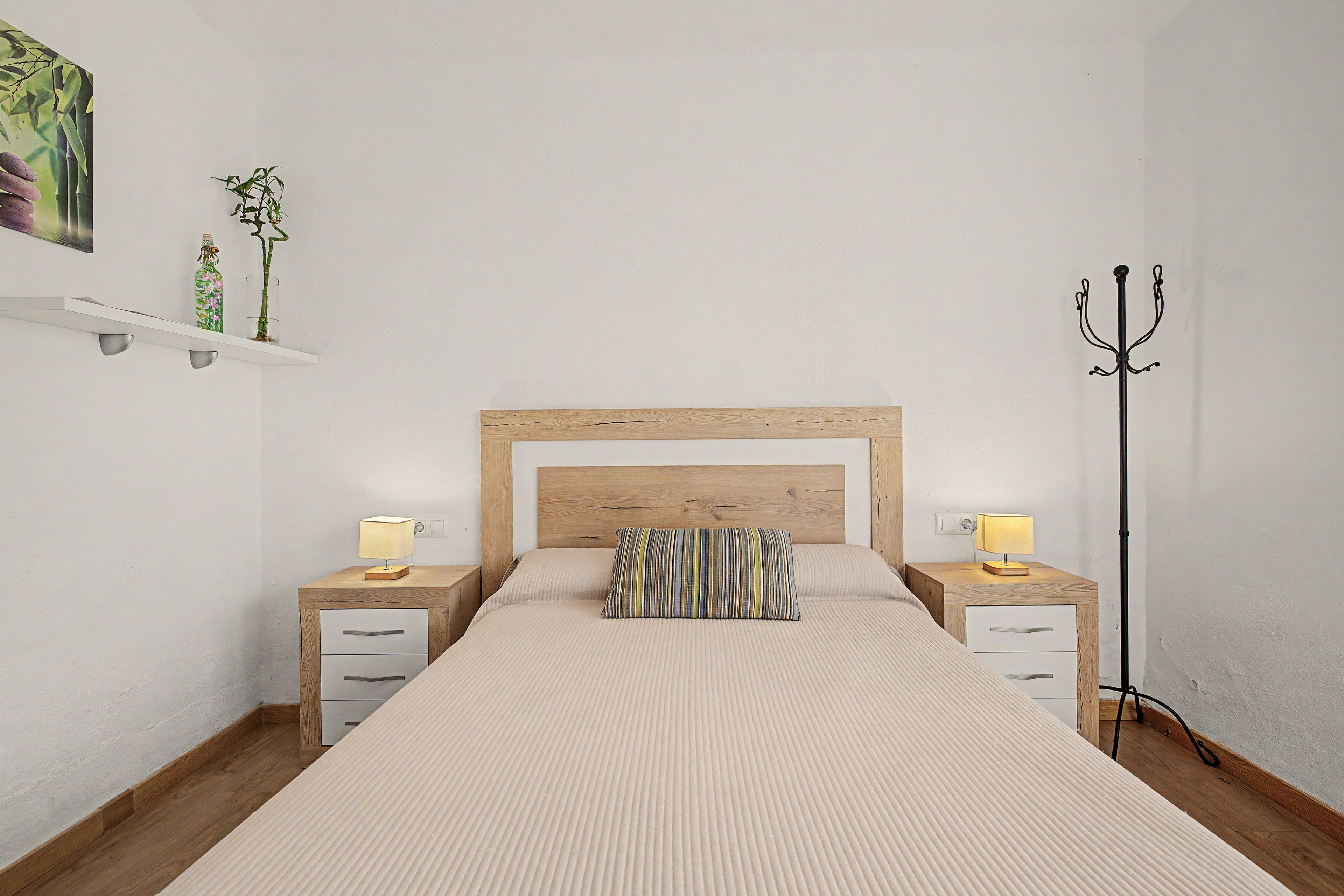 Un dormitorio ordenado con cabecera de madera, dos mesitas de noche con lámparas, un cojín a rayas y una lámpara de pie.