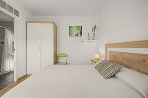 Un dormitori amb un llit, una cadira, una tauleta de nit, una prestatgeria amb objectes decoratius i una porta.