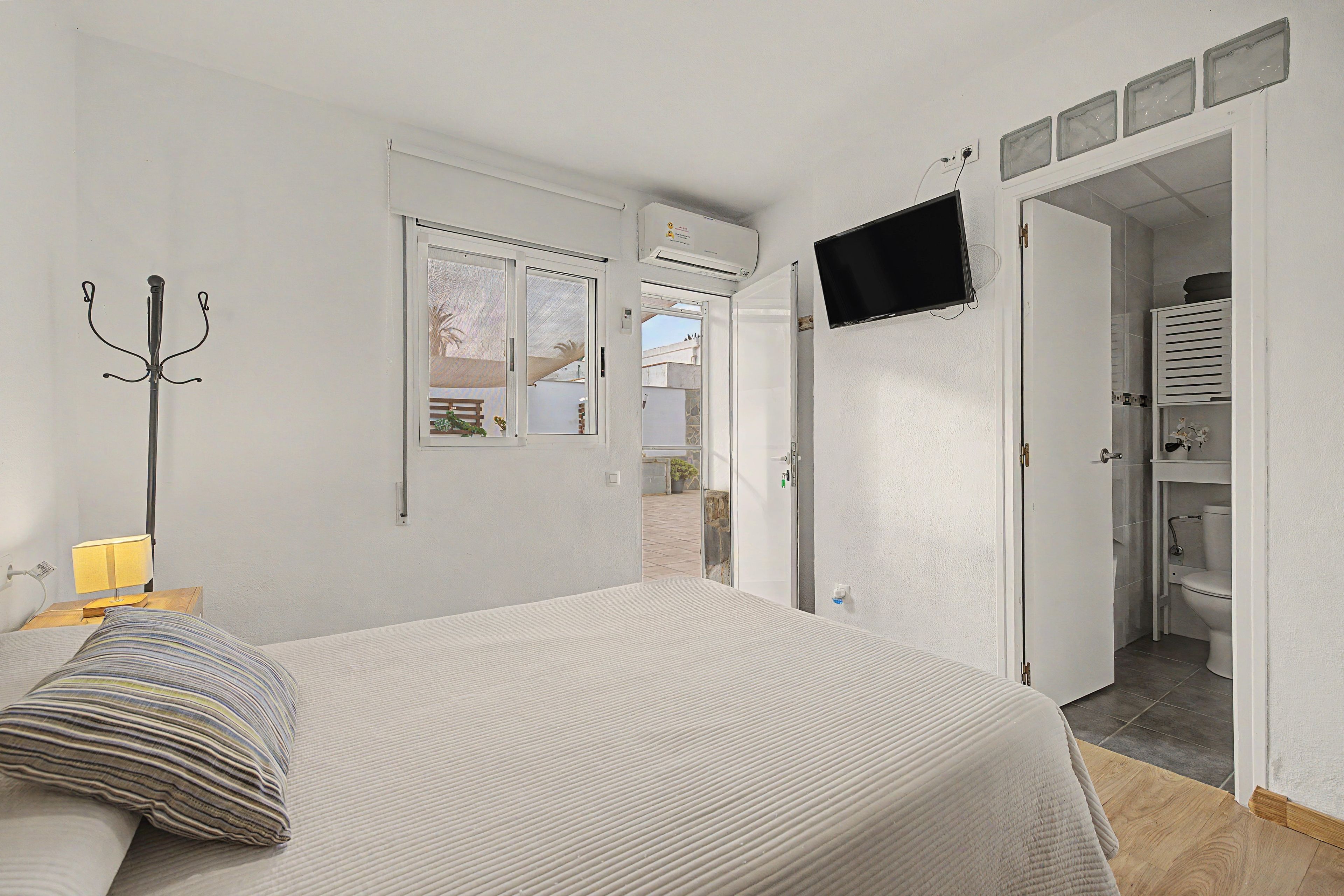 Un dormitorio con cama, mesita de noche con lámpara, perchero y televisor montado en la pared.