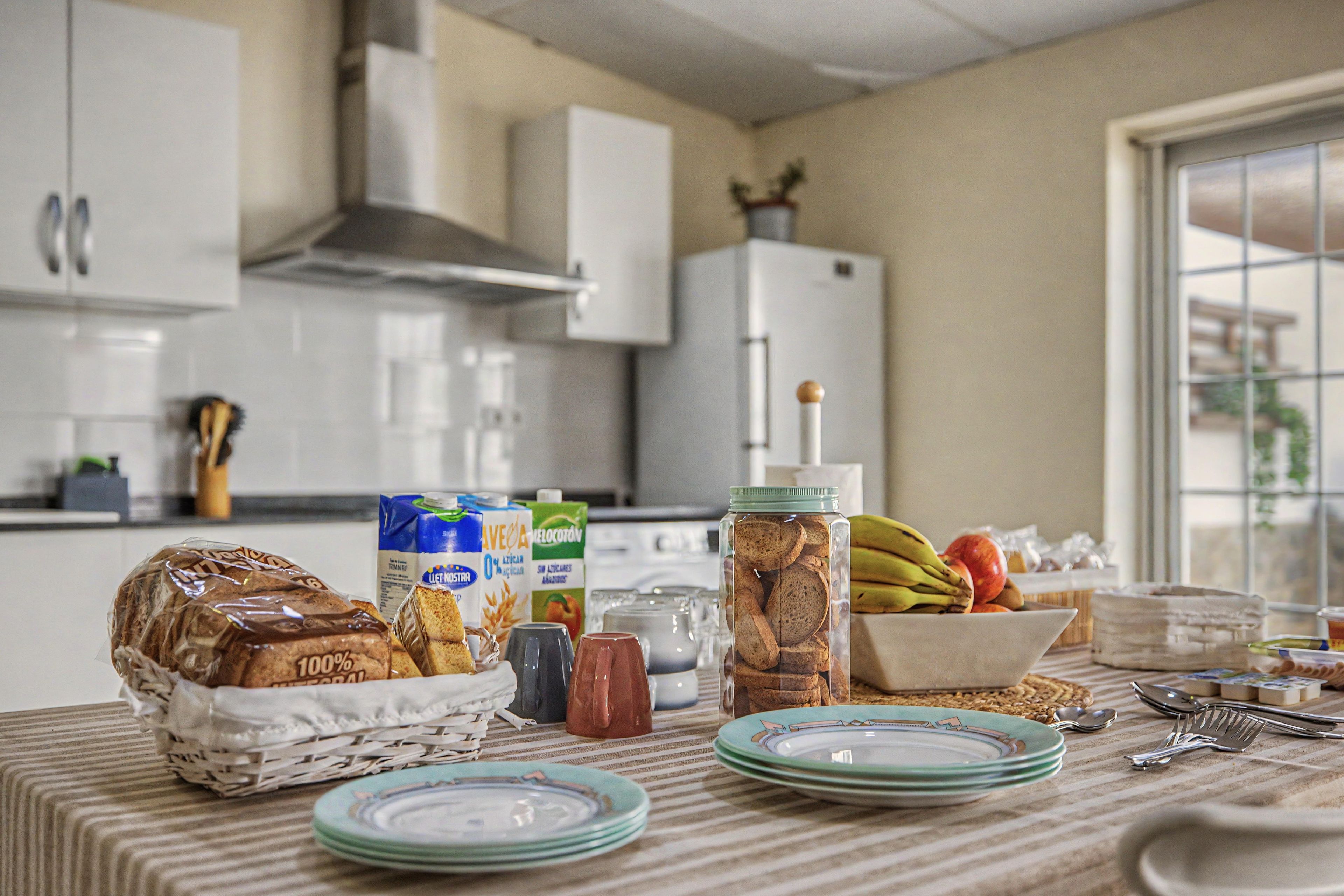 Una cocina con una mesa dispuesta para una comida, incluyendo pan, leche y un bol de fruta.