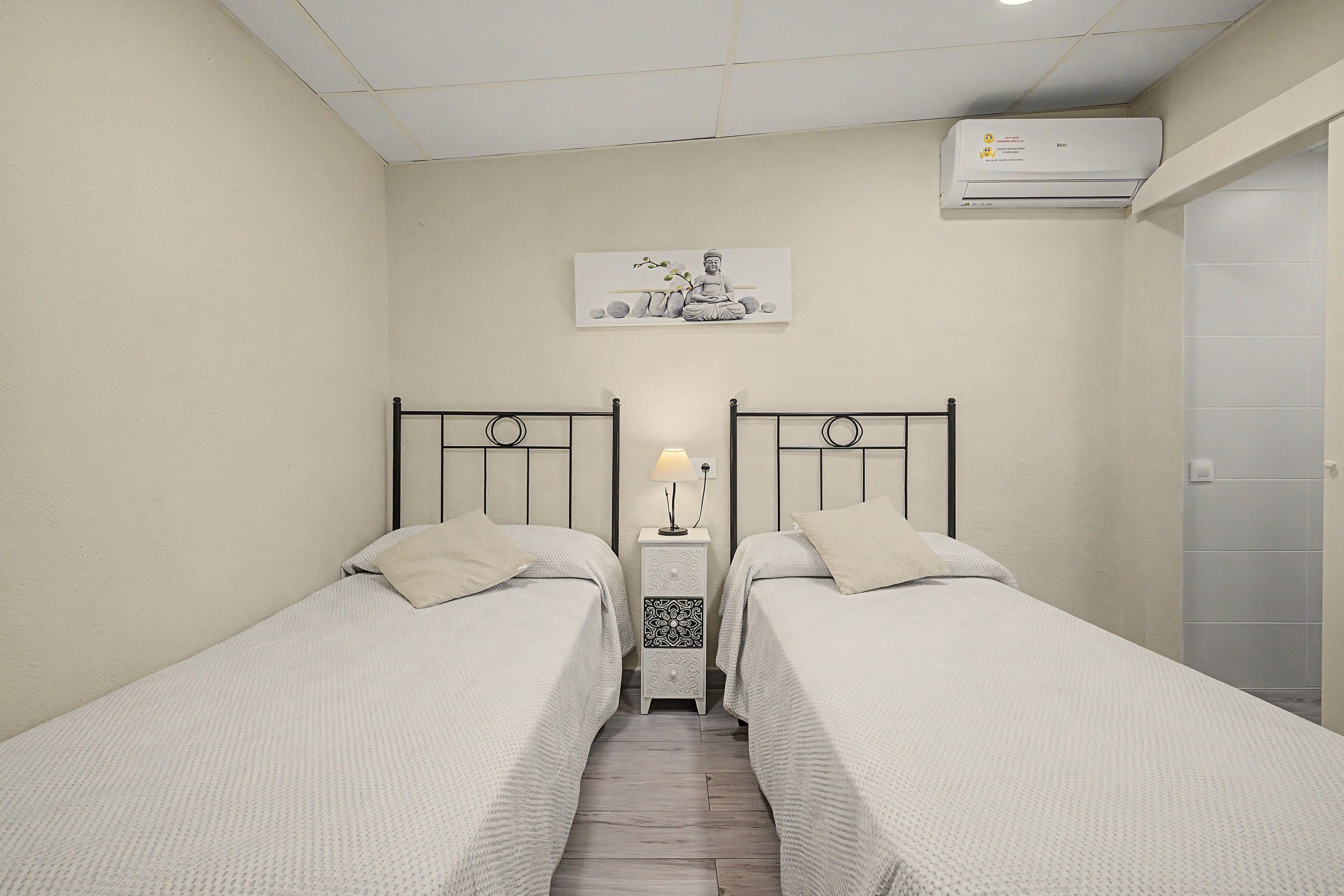 Habitación pequeña y limpia con dos camas individuales, una mesita de noche con lámpara, aire acondicionado de pared y un cuadro enmarcado sobre las camas.