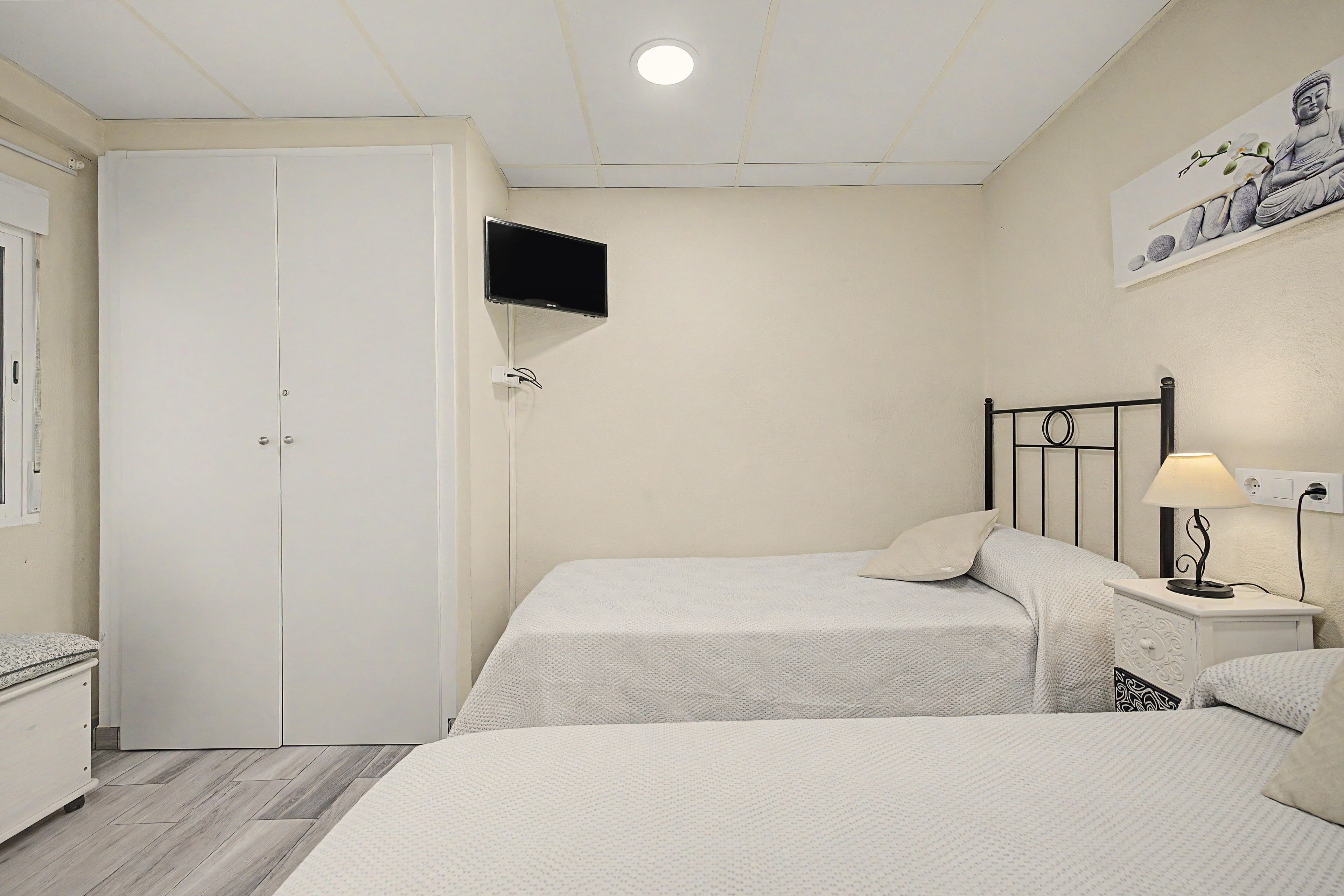 Un dormitorio con dos camas, televisión montada en la pared, mesita de noche con lámpara y armario.