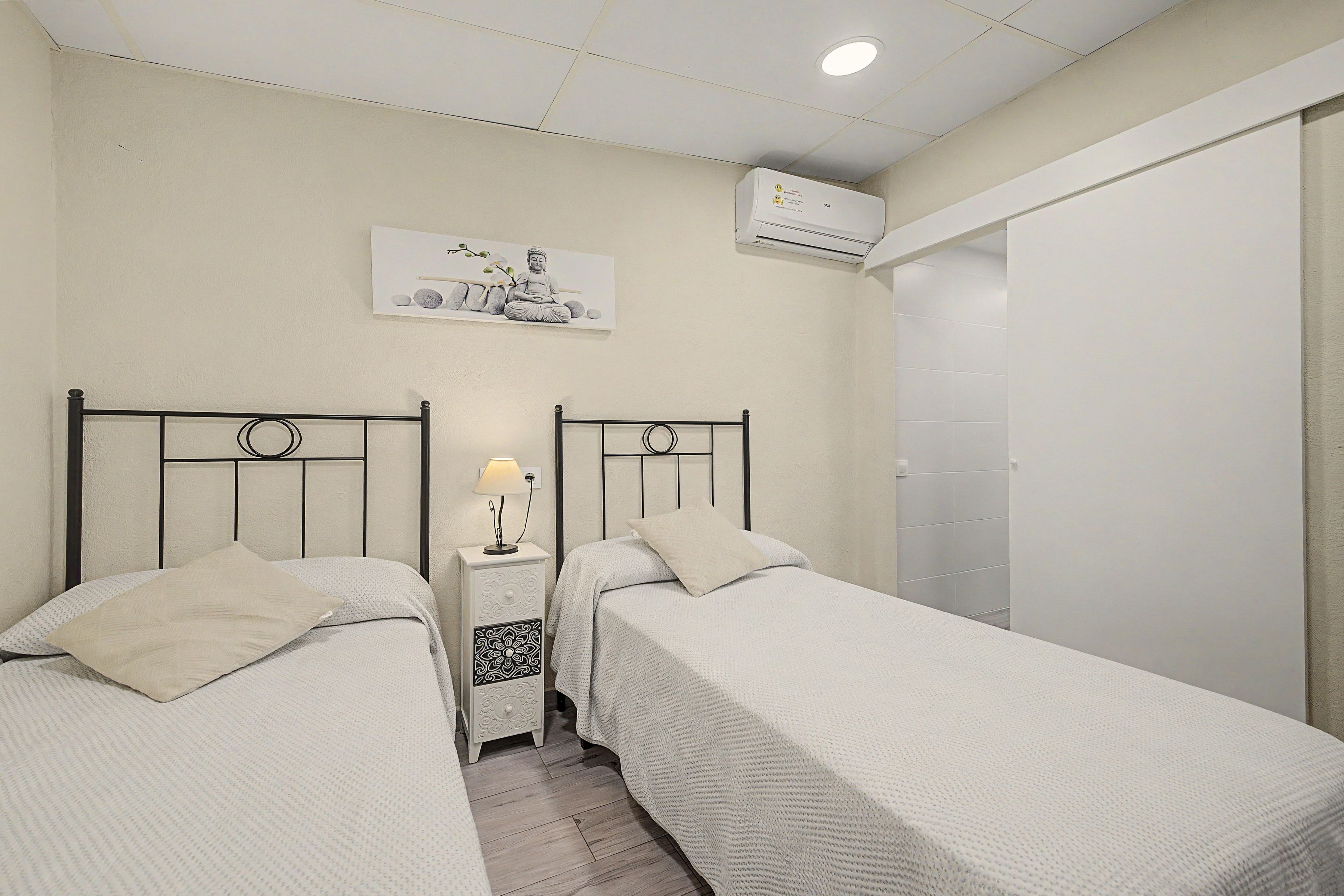 Une chambre avec deux lits simples, une table de chevet avec une lampe, un climatiseur mural et un tableau au-dessus des lits.