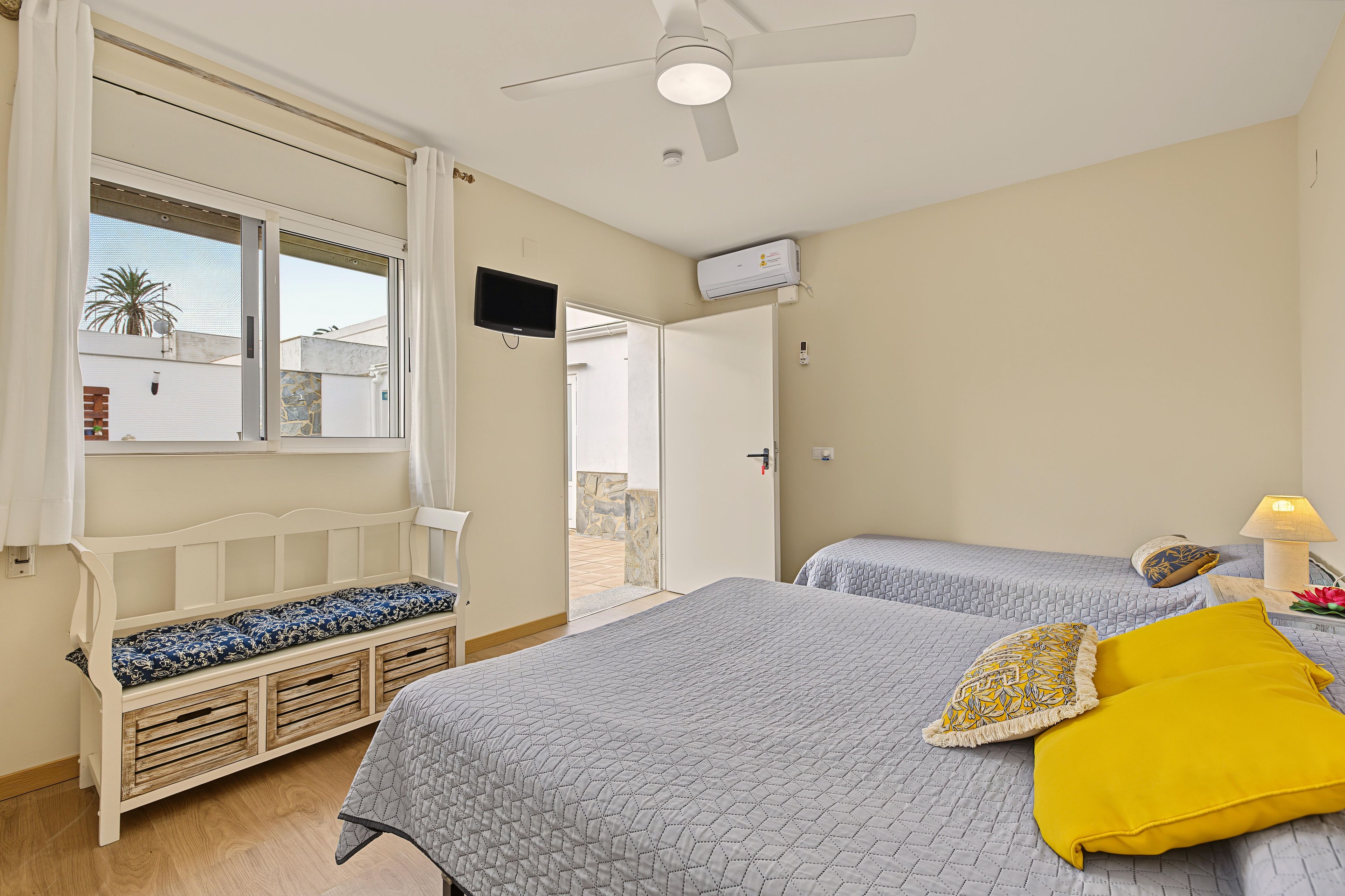 Une chambre avec deux lits, un ventilateur de plafond, une télévision et une fenêtre donnant sur un immeuble et des palmiers.