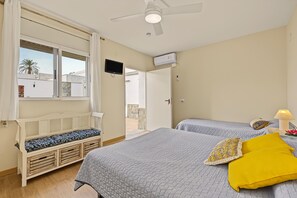 Une chambre avec deux lits, un ventilateur de plafond, une télévision et une fenêtre donnant sur un immeuble et des palmiers.