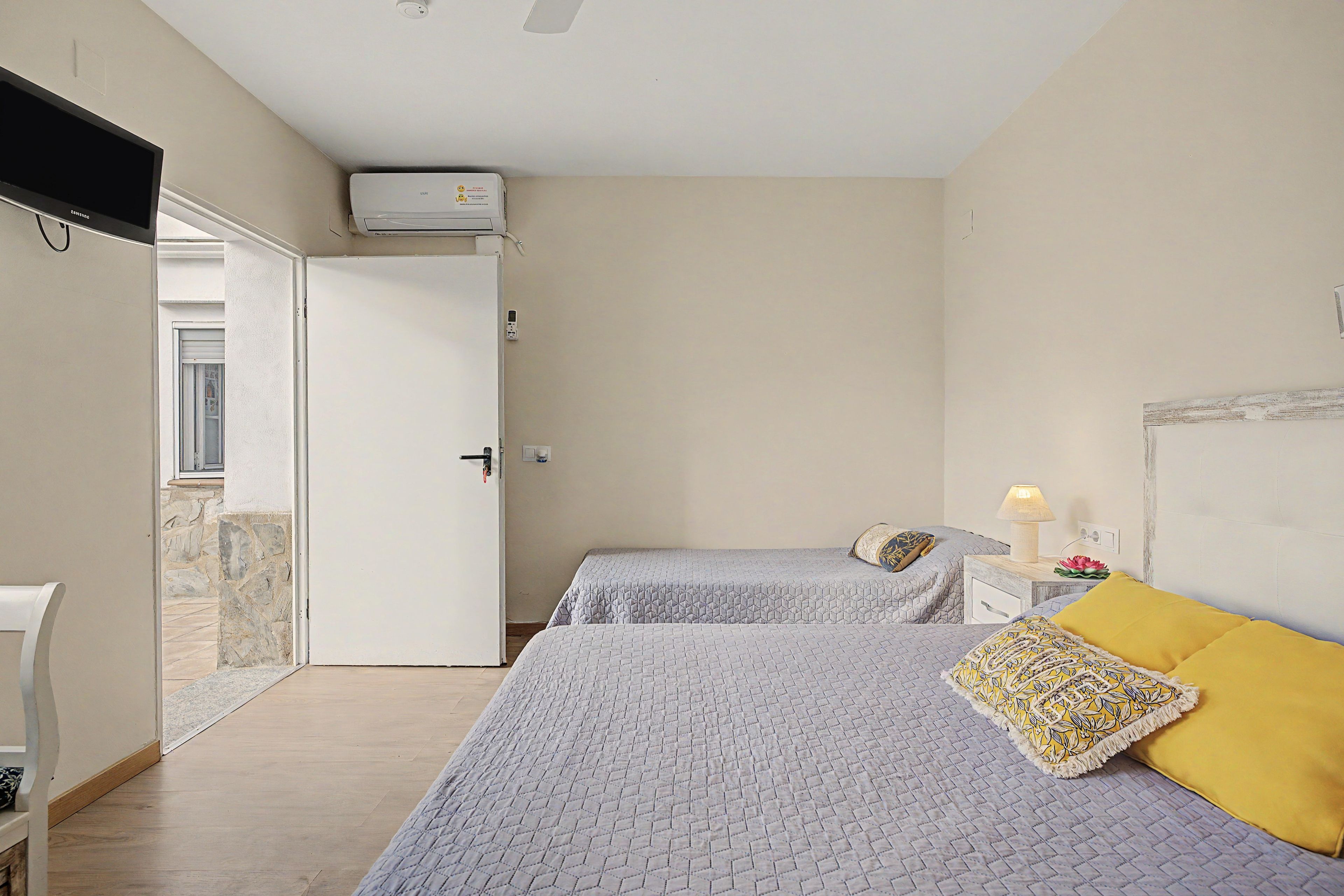 Een slaapkamer met twee bedden, een witte deur, een klein nachtkastje met een lamp en een aan de muur bevestigde airconditioner.