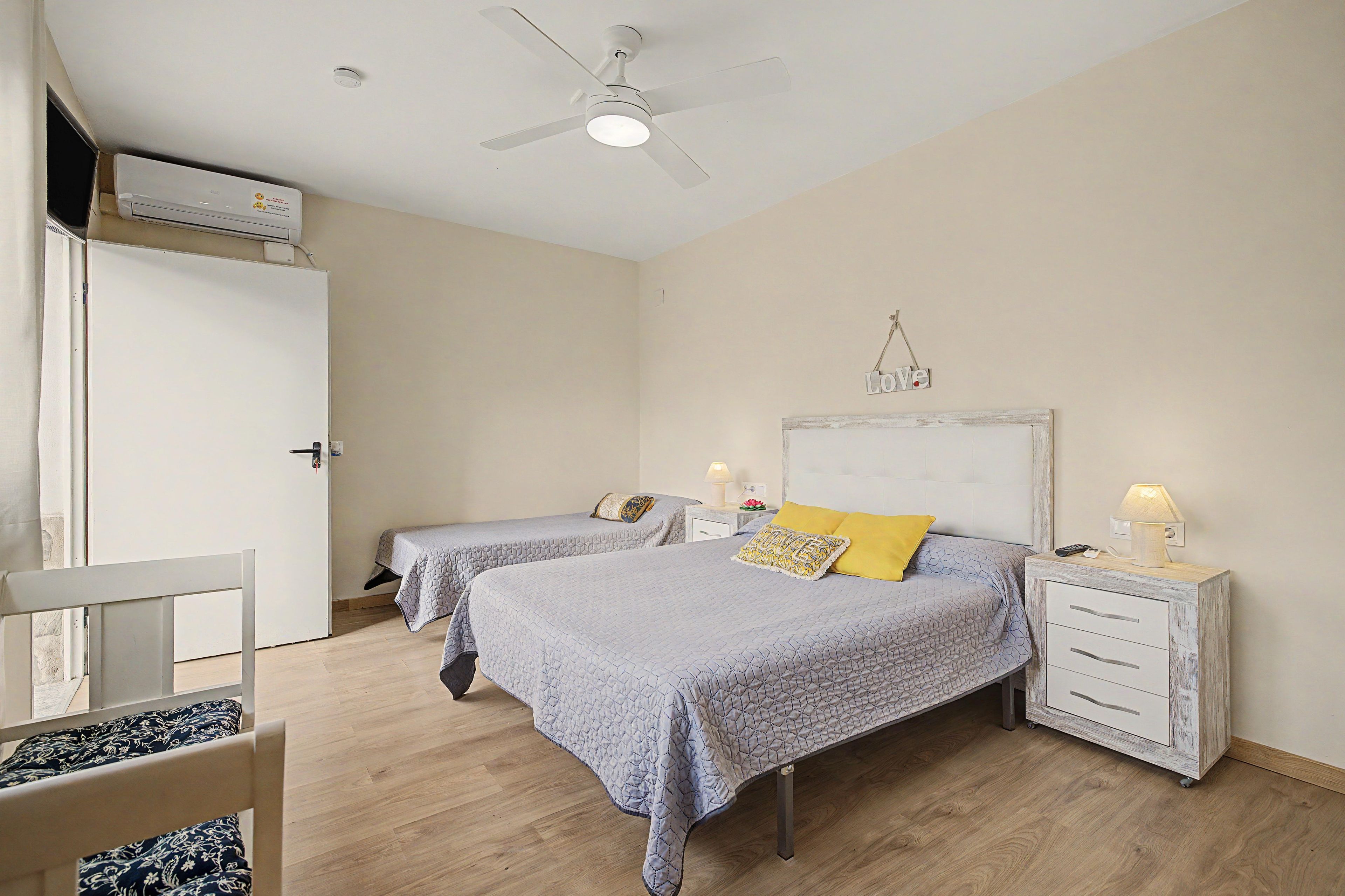 A bedroom with two beds, a ceiling fan, a bedside table with a lamp, and a wall-mounted light fixture.